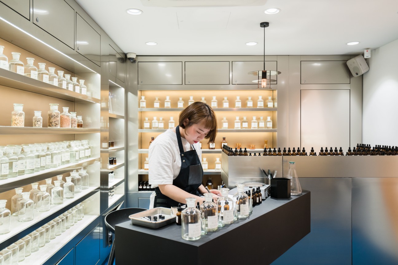 A staff member works at a counter in Gentle Monster's scent-themed shop, surrounded by glass vials and bottles displayed on illuminated shelves.
