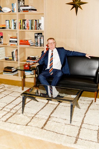 Theatre agent George Lane sits on a black leather sofa in his office, wearing a navy suit and striped tie.