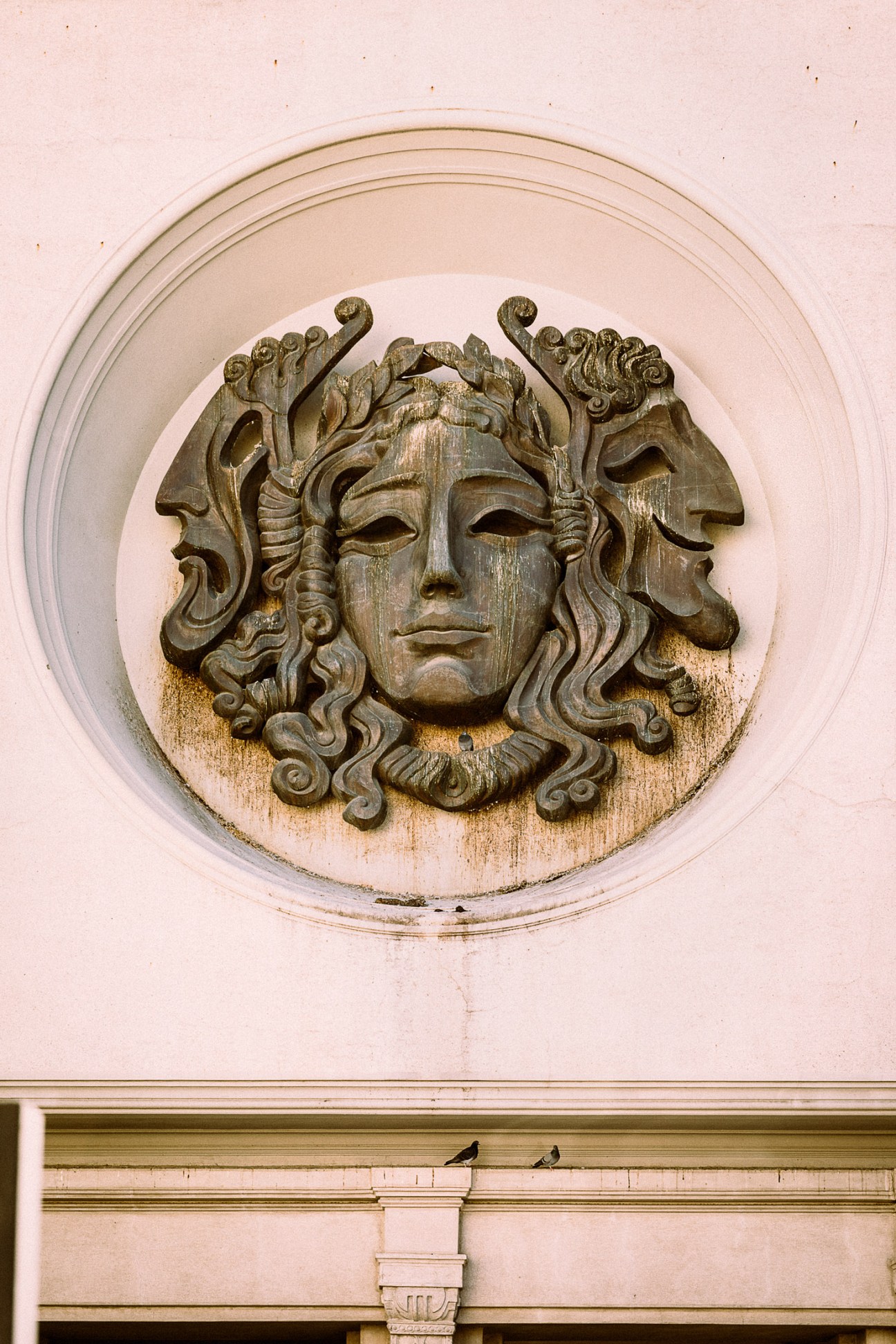 Ornate stone relief sculpture of a classical face with elaborate curled hair and two profile heads on either side on a pink building facade.