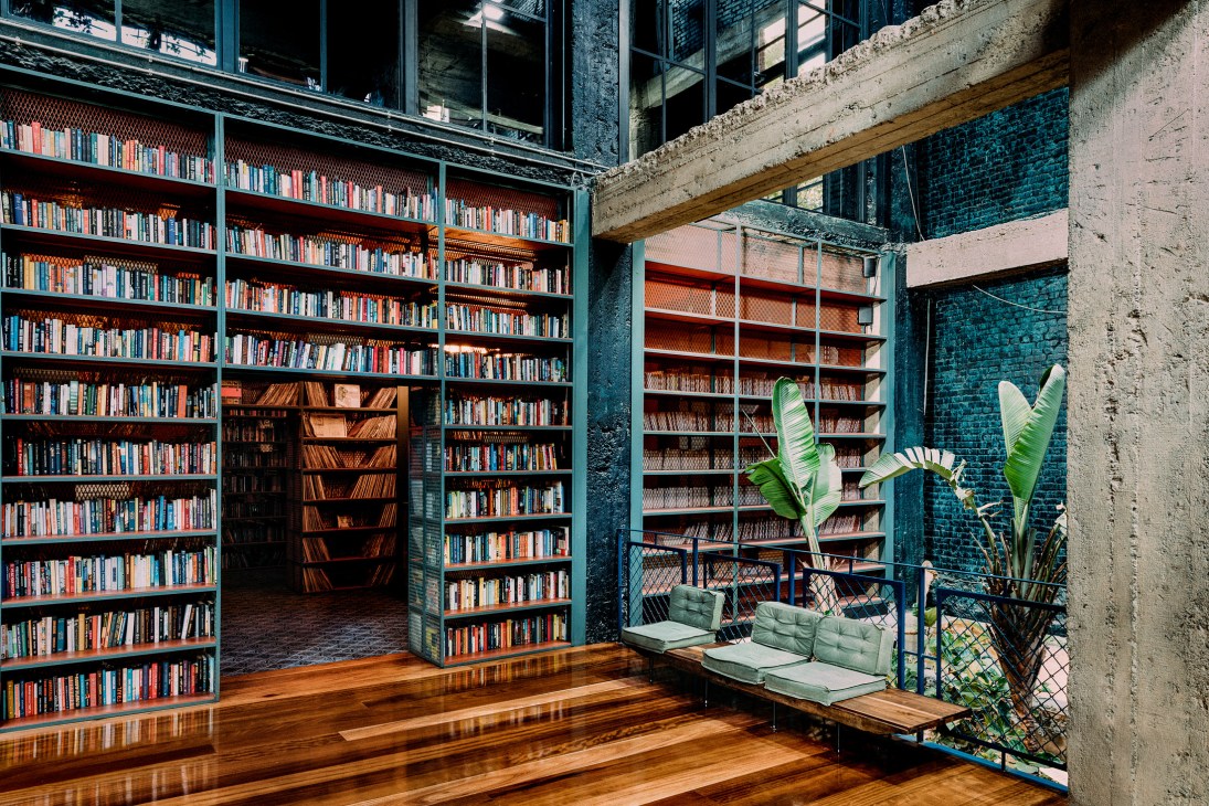 Industrial courtyard with bookshelves, green cushioned seating, and tropical plants at Stamba Hotel.