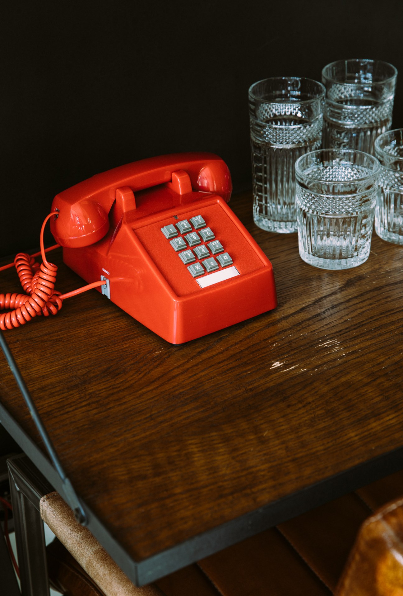 Red rotary telephone with coiled cord on wooden table next to crystal glasses