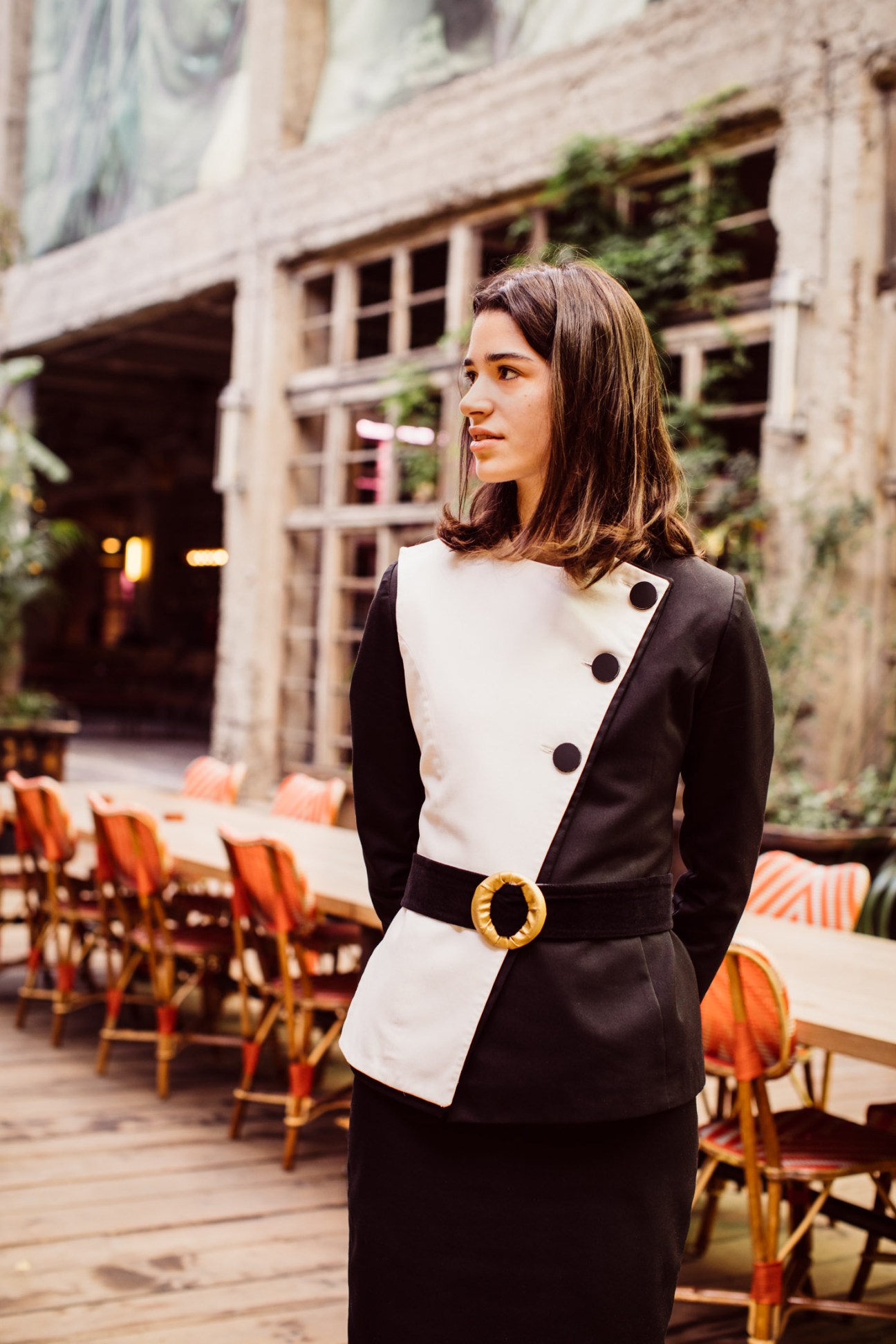 Woman in black and white blazer with gold belt standing in Soviet-era courtyard with orange chairs.