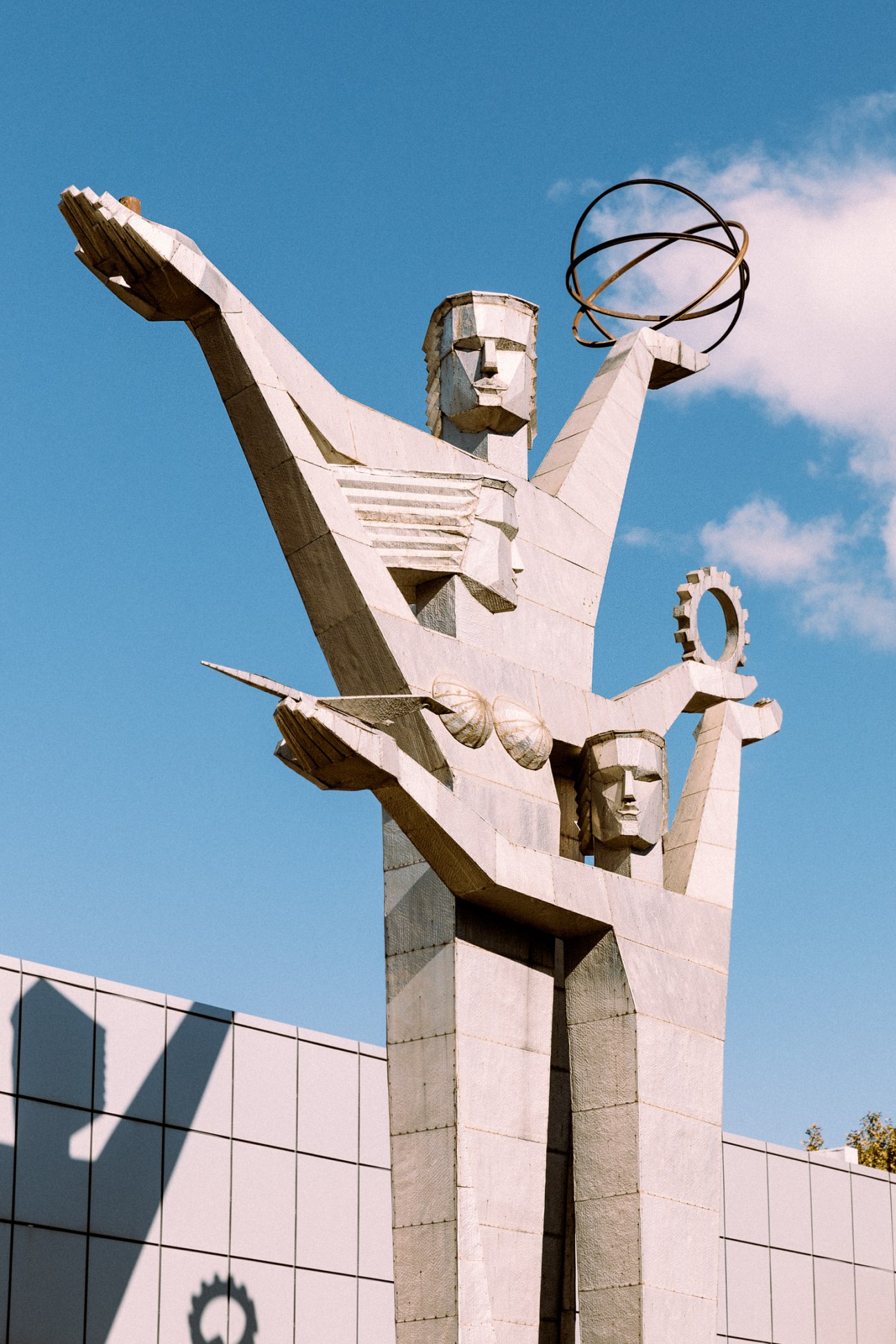 Monumental white stone sculpture of figures with raised arms holding spherical objects against blue sky