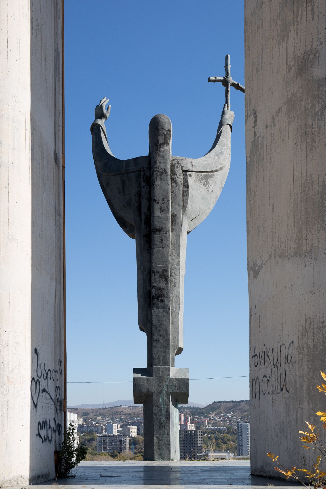 Stone statue of Saint Nino holding a cross, viewed through concrete columns against blue sky
