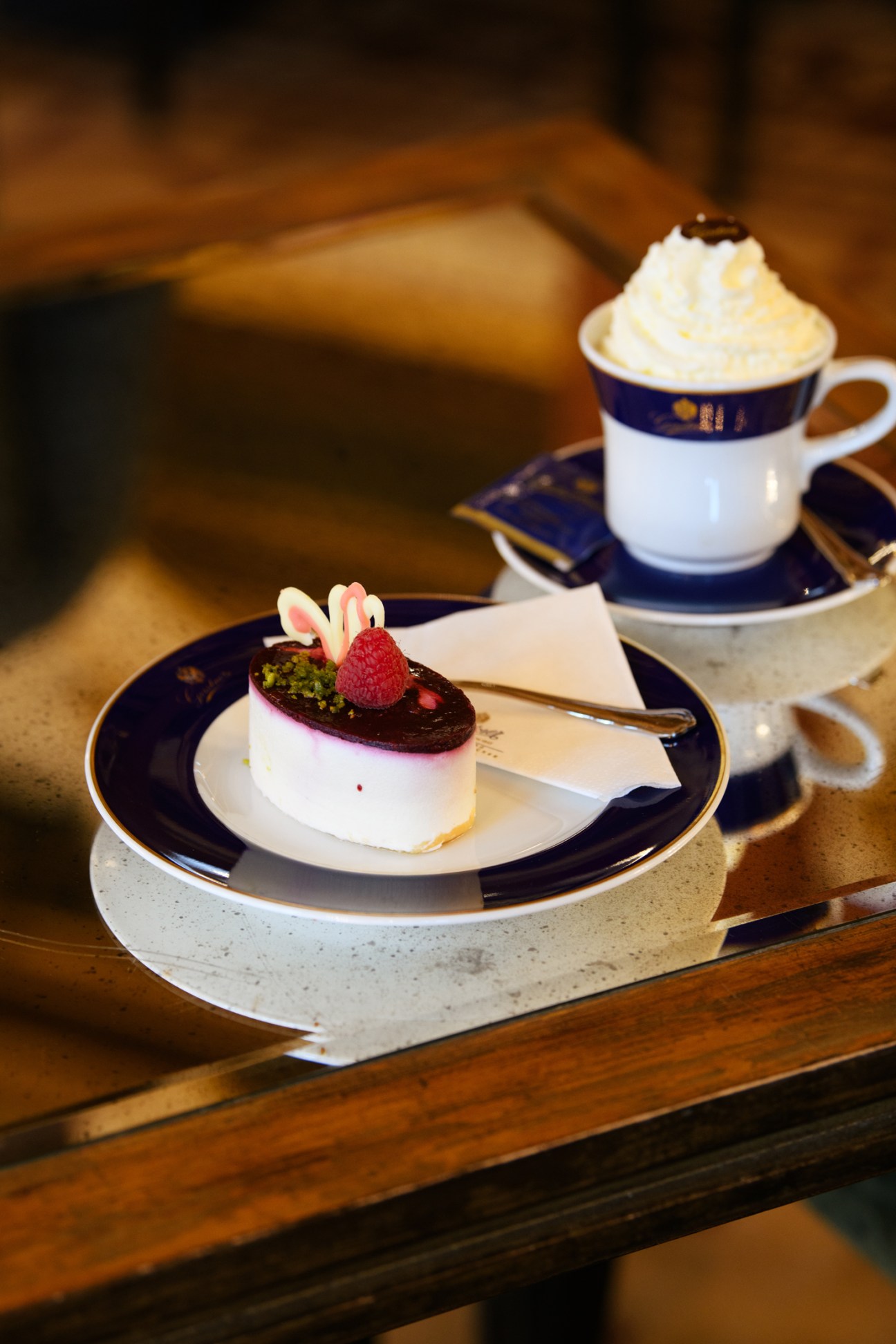 White cheesecake with chocolate glaze, raspberry and garnish on blue-rimmed plate with coffee cup