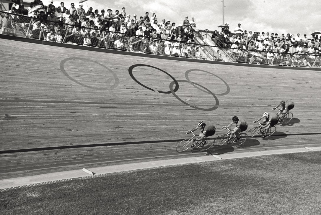 Black and white photograph of cyclists racing on a velodrome track with Olympic rings painted on the wooden surface and spectators in the stands.