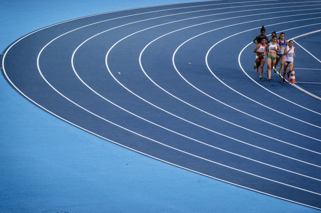 Female track athletes running on a bright blue Olympic track with white lane markings.