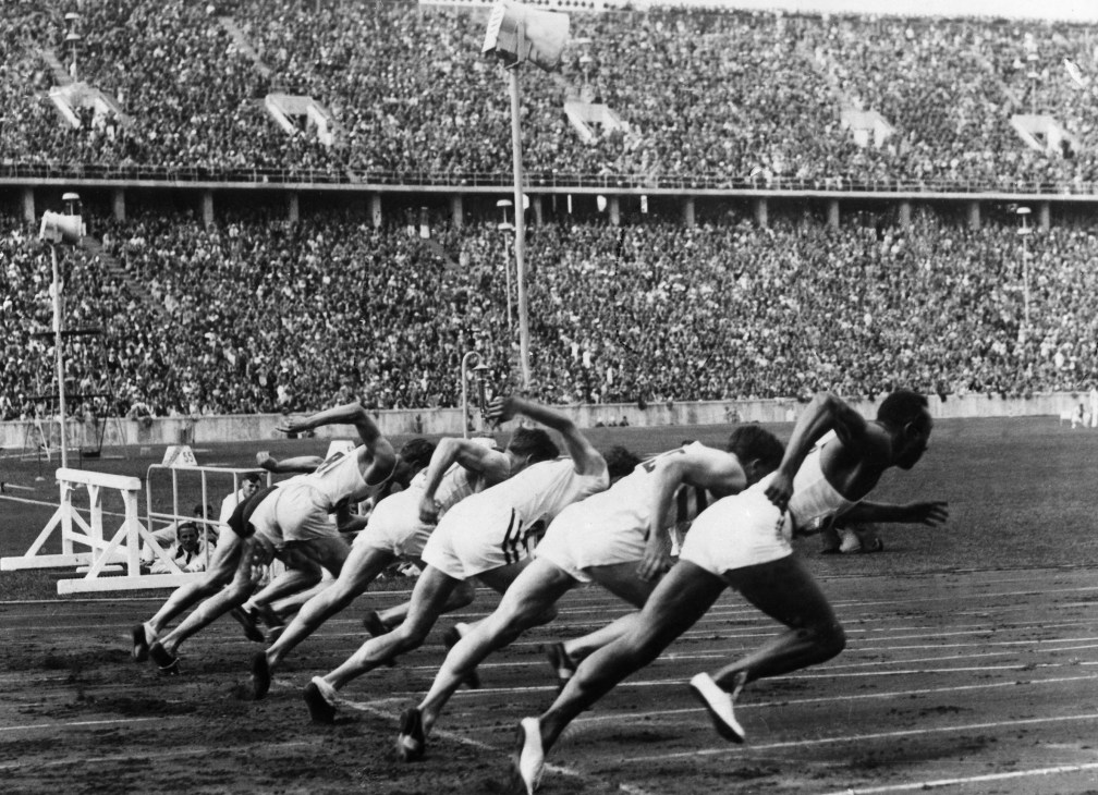Black and white photograph of sprinters at the starting line during the 1936 Berlin Olympics with a packed stadium in the background.