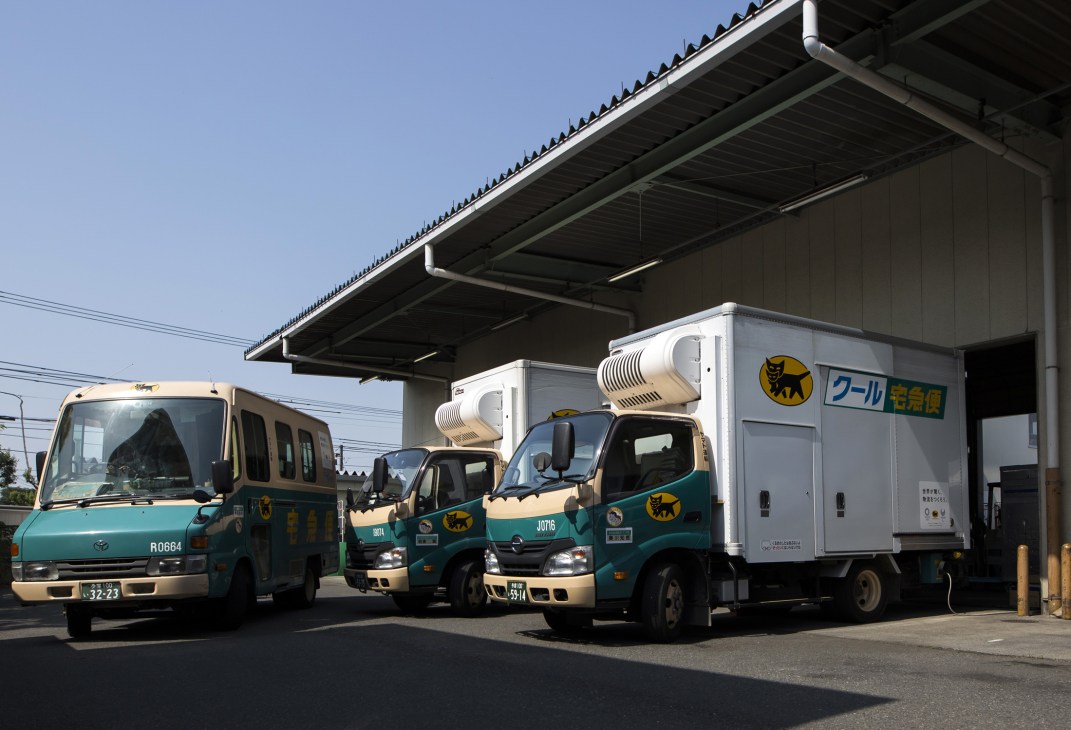 Ta-Q-Bin delivery fleet parked under an overpass, featuring green-and-tan Hino and other branded trucks with roof-mounted cooling units.