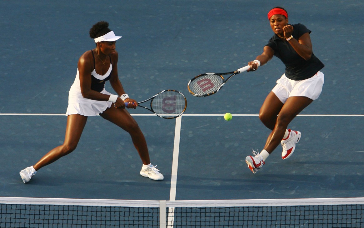 The Williams sisters playing doubles tennis on a blue court.