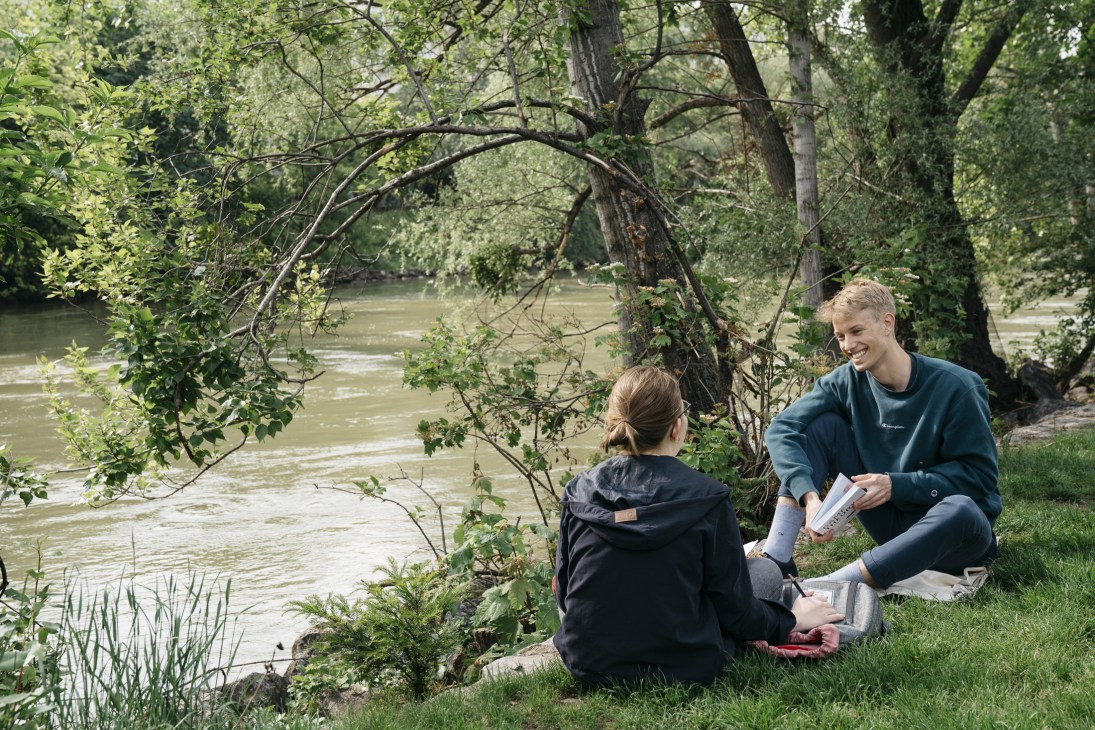 Two people sit on grass by a river under overhanging tree branches, looking at reading material together.