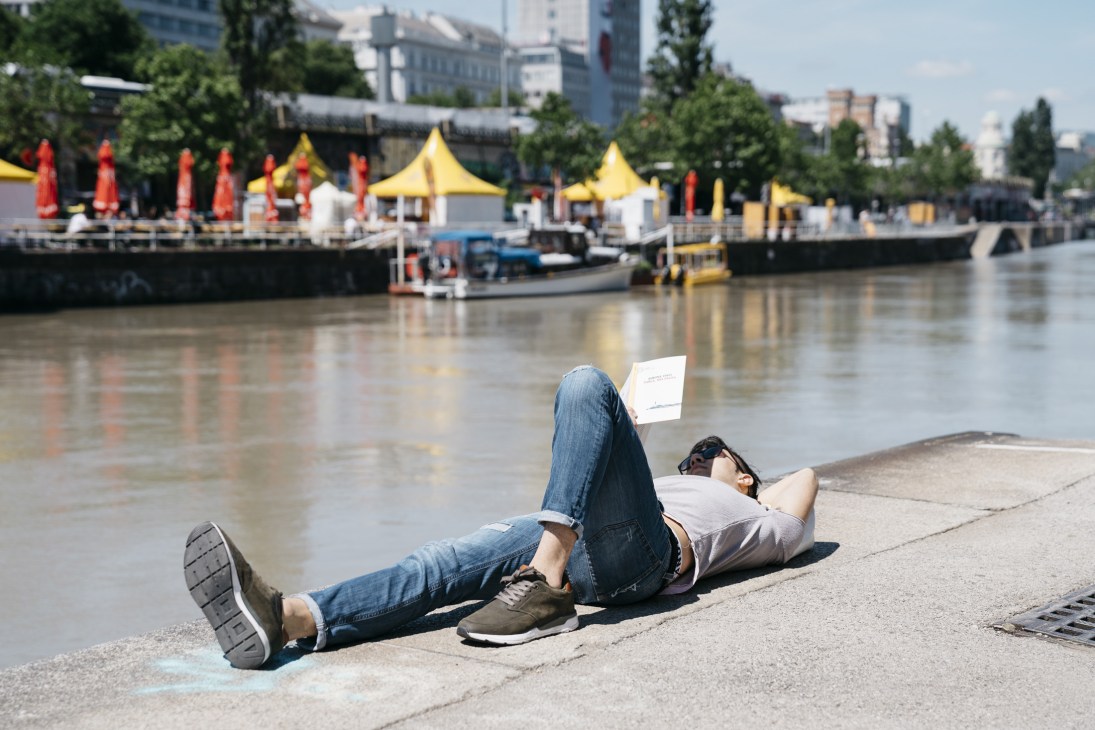 Person lying on concrete waterside promenade reading, with Danube Canal bars and yellow umbrellas blurred in background.