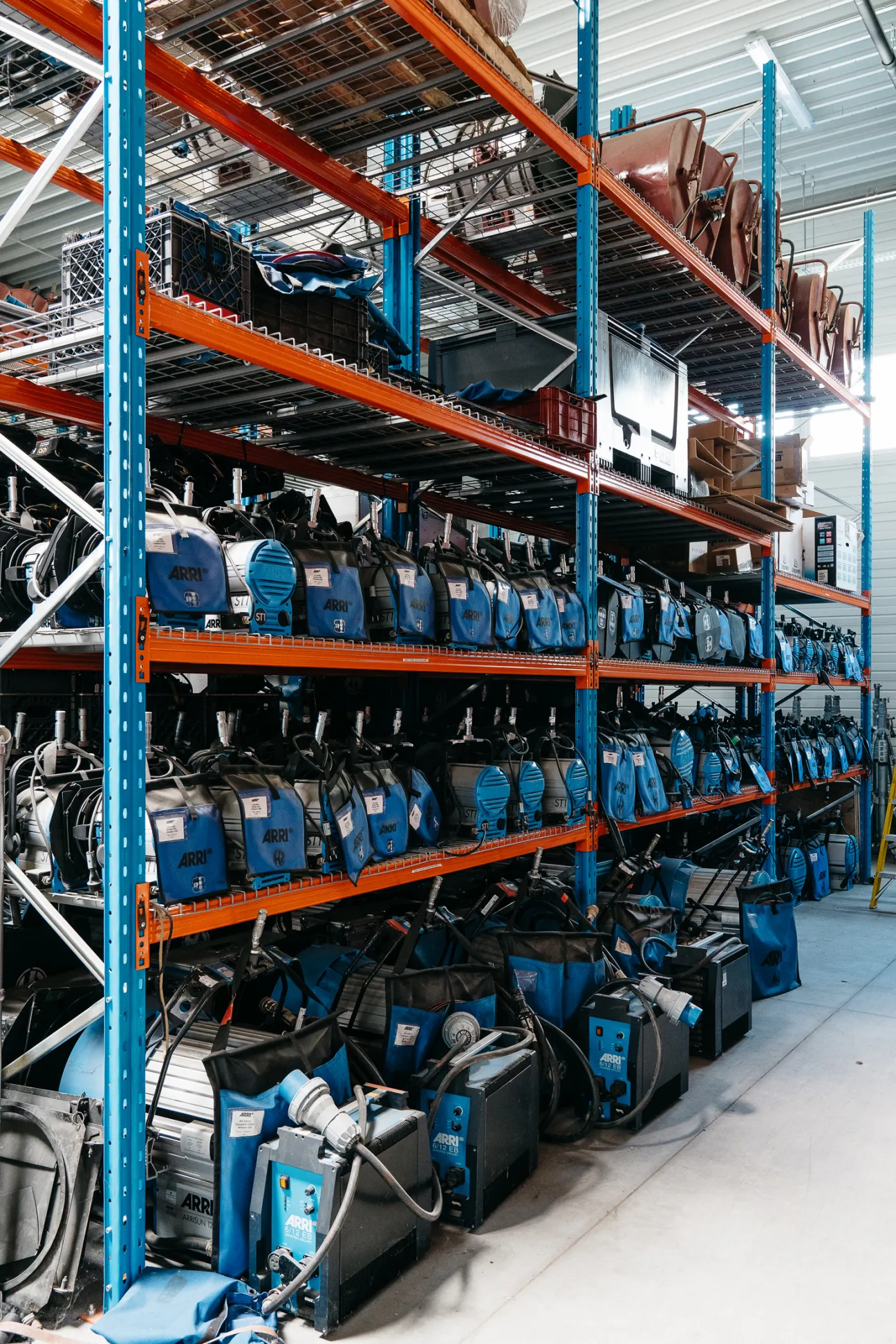 Industrial shelving with blue ARRI lighting and grip equipment stored on multiple levels in a warehouse.