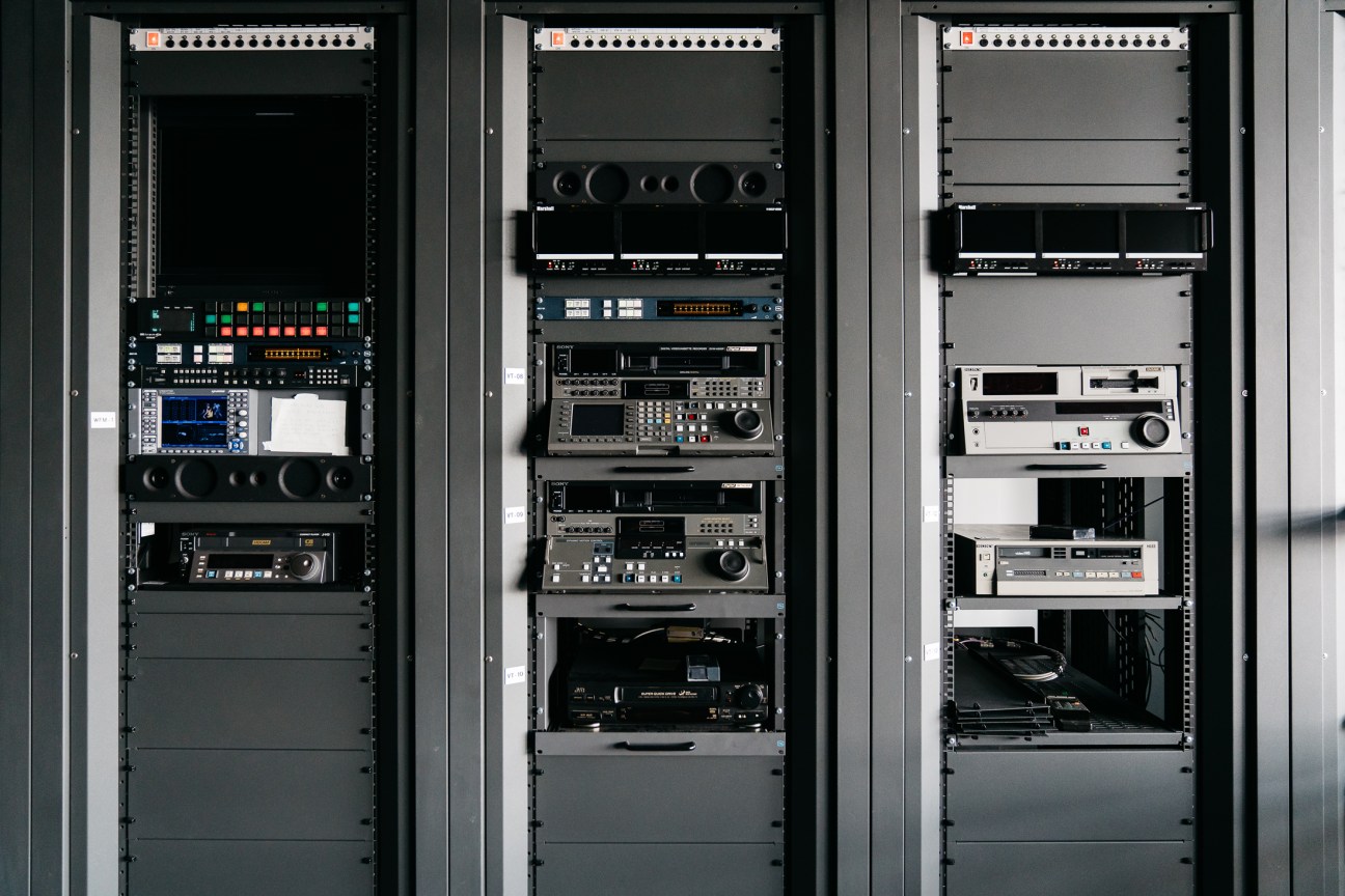 Three tall equipment racks with professional video and audio editing hardware and controls in a post-production facility.