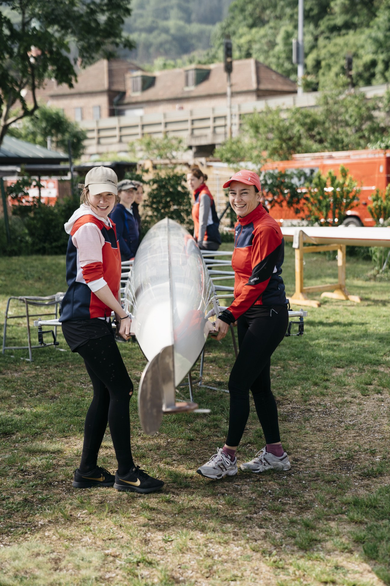 Two people in red and navy athletic wear carry a rowing boat between them at a riverside facility.