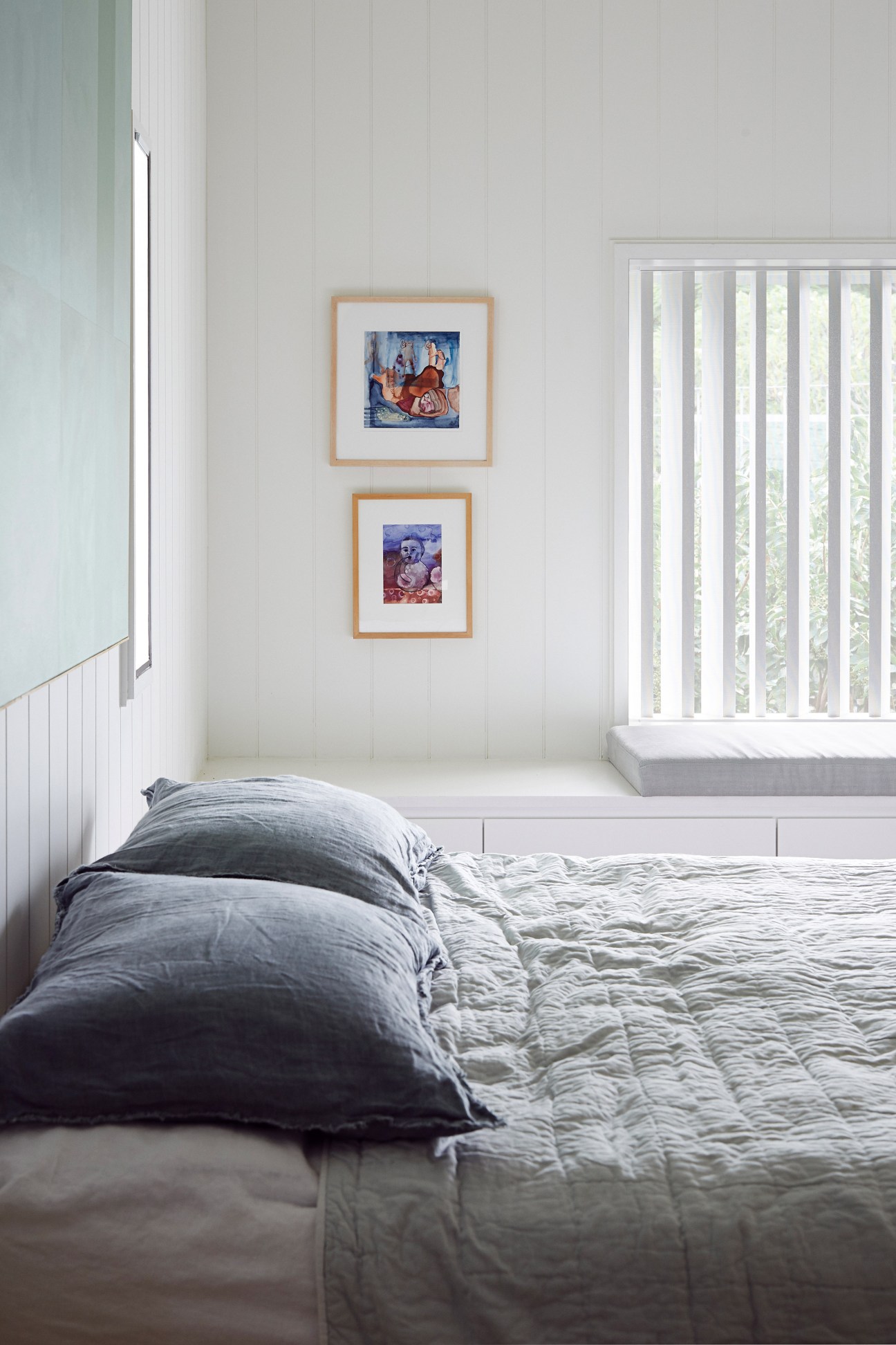 Minimalist bedroom with white walls, bed with grey bedding, two framed artworks, and vertical blinds on window.