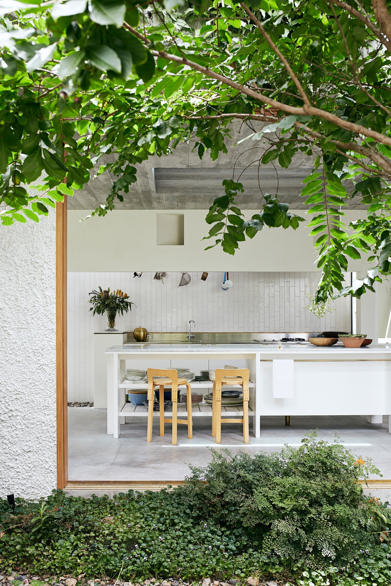 Kitchen with white subway tile backsplash, marble countertop, brass rail, and wooden chairs, framed by hanging greenery.