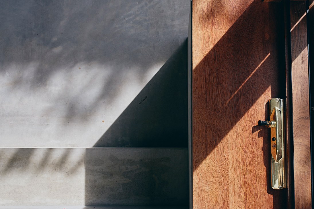 Wooden door with brass hardware casting shadows in sunlight against concrete wall.