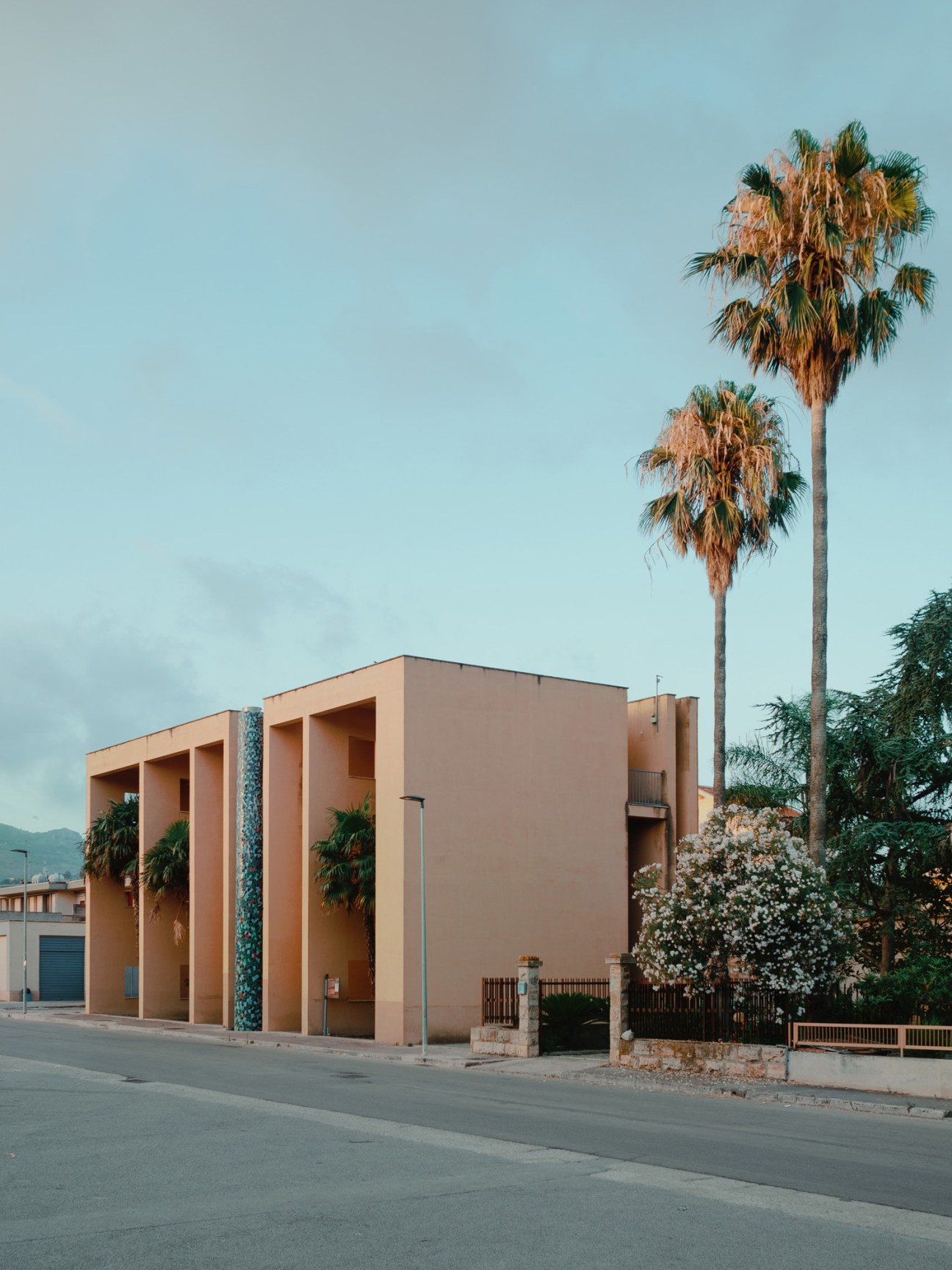 Terracotta-colored modernist building with deep archways and tall palm trees behind, designed by Franco Purini and Laura Thermes.