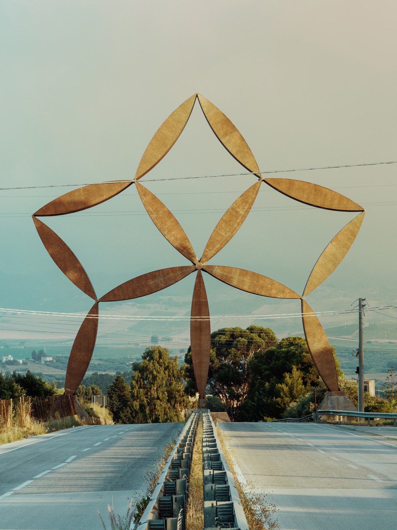 Large abstract steel sculpture with star-like form spanning across a road in Sicily.