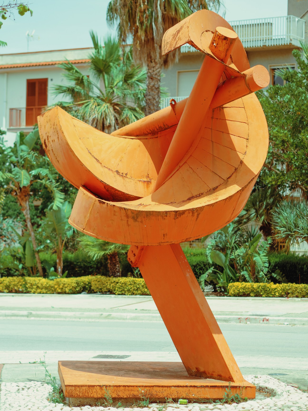 Spiral-shaped concrete sculpture in outdoor setting with palm trees and Mediterranean buildings.