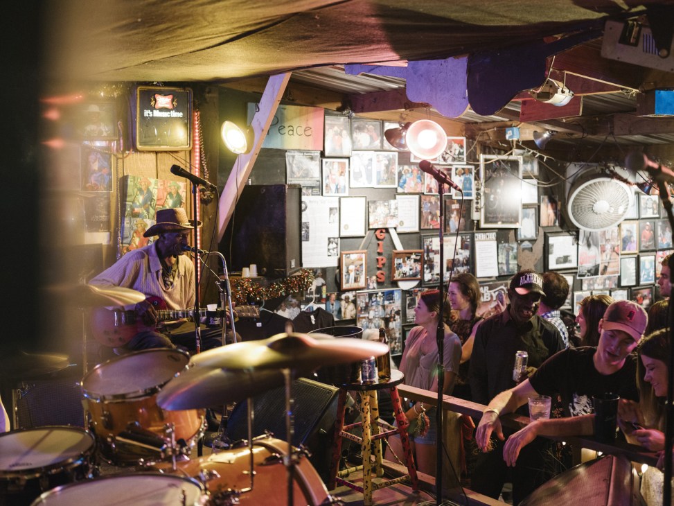 Gip Gipson performs with guitar at his juke joint, surrounded by patrons and memorabilia on the walls.