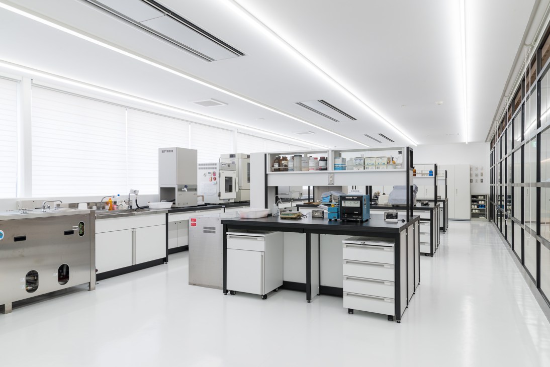 Modern laboratory with white linear ceiling lights, scientific testing equipment, workbenches, and storage systems for textile development and quality control.