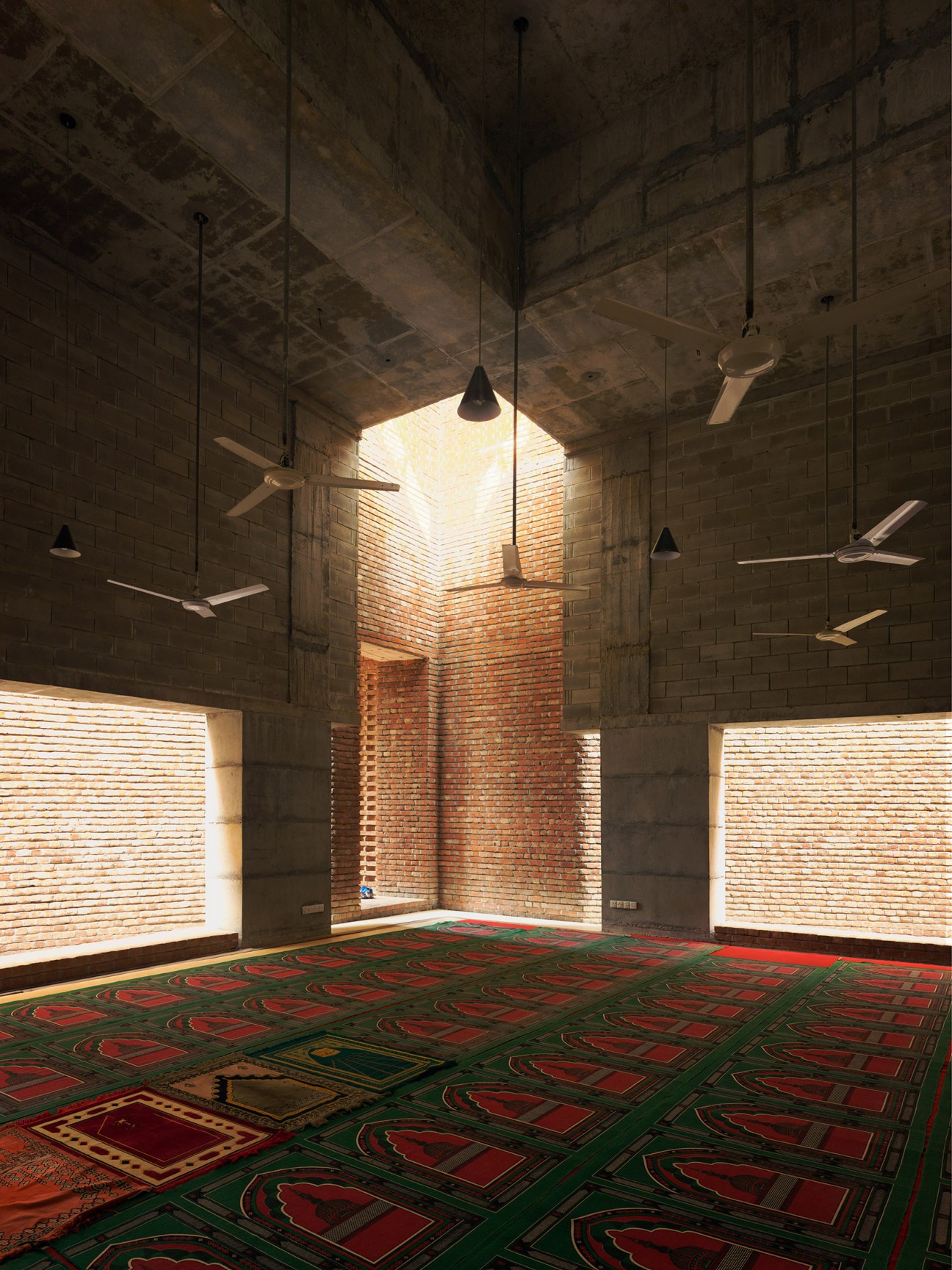 Interior of Bait Ur Rouf Mosque with brick walls, hanging ceiling fans, natural light streaming through windows, and decorative prayer carpet on floor.