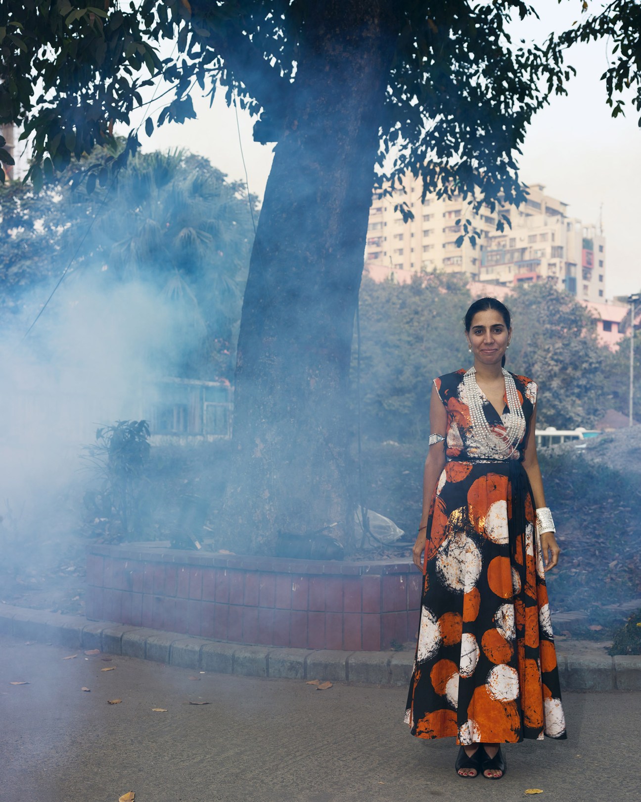 Diana Campbell Betancourt stands on a Dhaka street beneath a large tree, with apartment buildings visible in hazy background.
