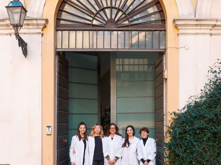 Five women in white lab coats standing in a doorway of a cream-colored Roman building with ivy.