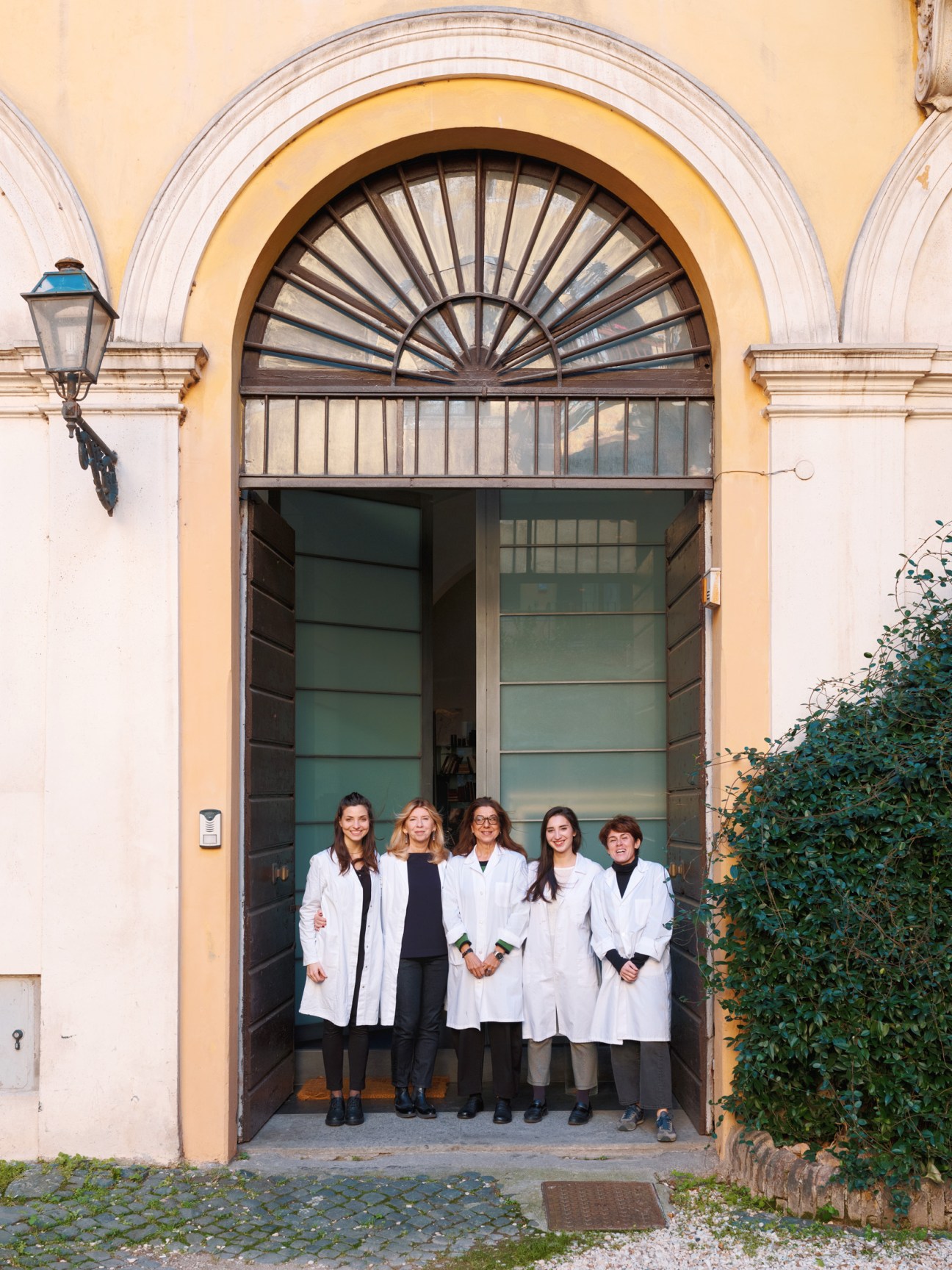 Five women in white lab coats standing in a doorway of a cream-colored Roman building with ivy.