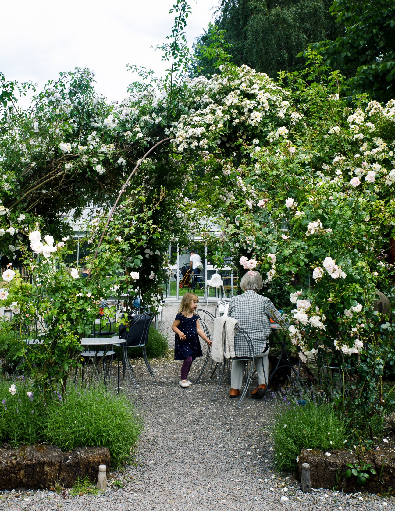 A child and elderly man sit at a table under a large flowering rose archway in a garden cafe