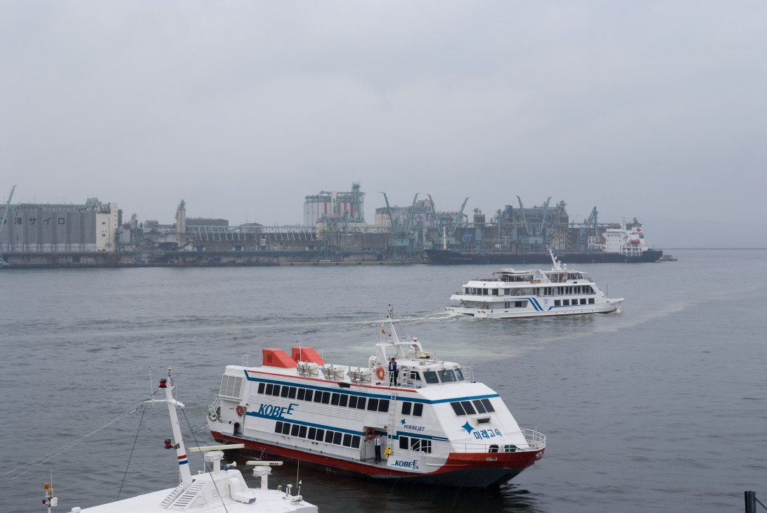 A white and red ferry boat named Kobe Mirari docked at Hakata Port with industrial port buildings visible in the background.