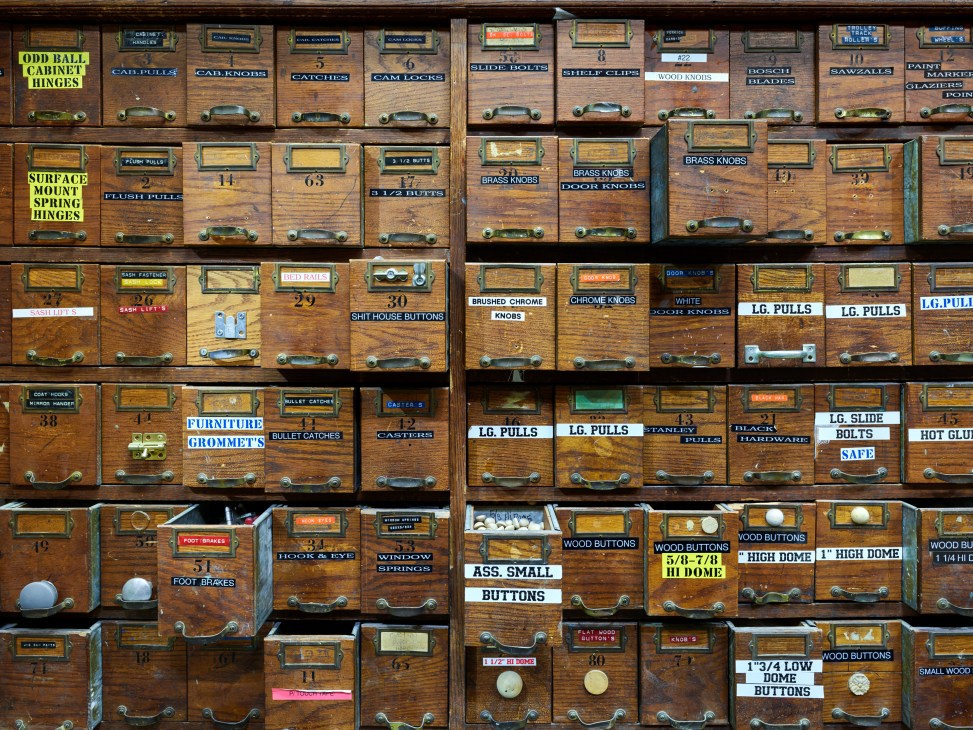 Wooden apothecary cabinet with numerous labeled drawers containing hardware components and buttons for set construction.