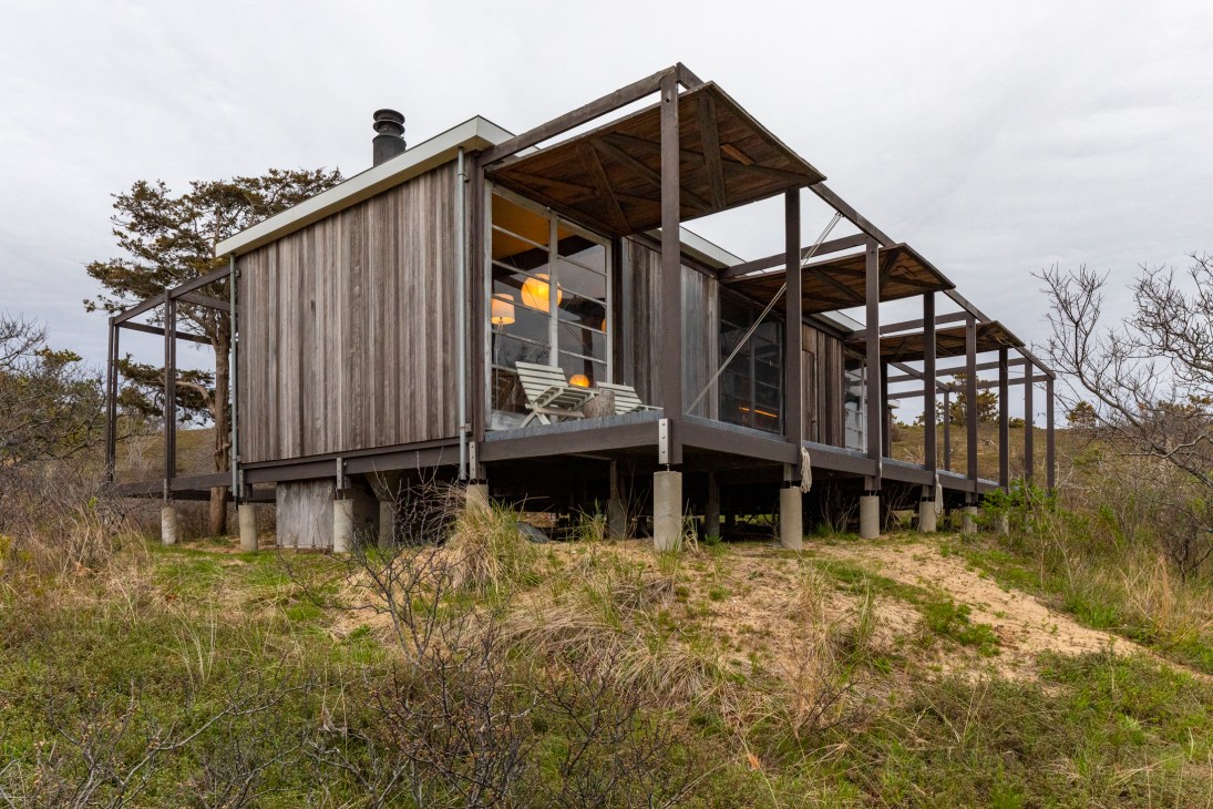 Modernist wooden house on stilts with open hatches and metal frames amid sandy dunes and sparse vegetation.