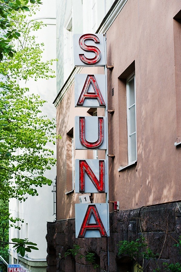 Vertical red neon sign reading 'SAUNA' mounted on a brick building with green foliage visible.
