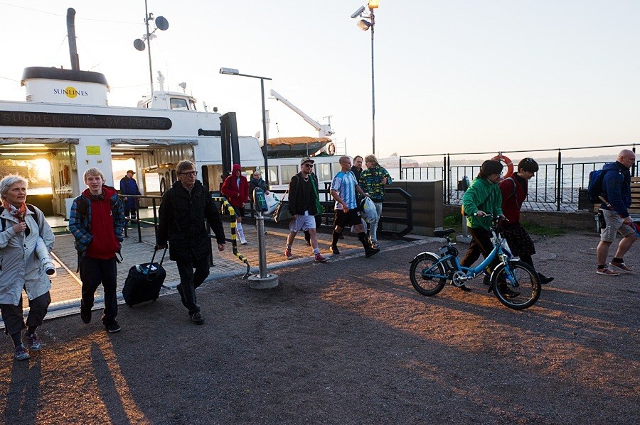 Ferry terminal with passengers, bicycles, and a child on a bike near a docked vessel.