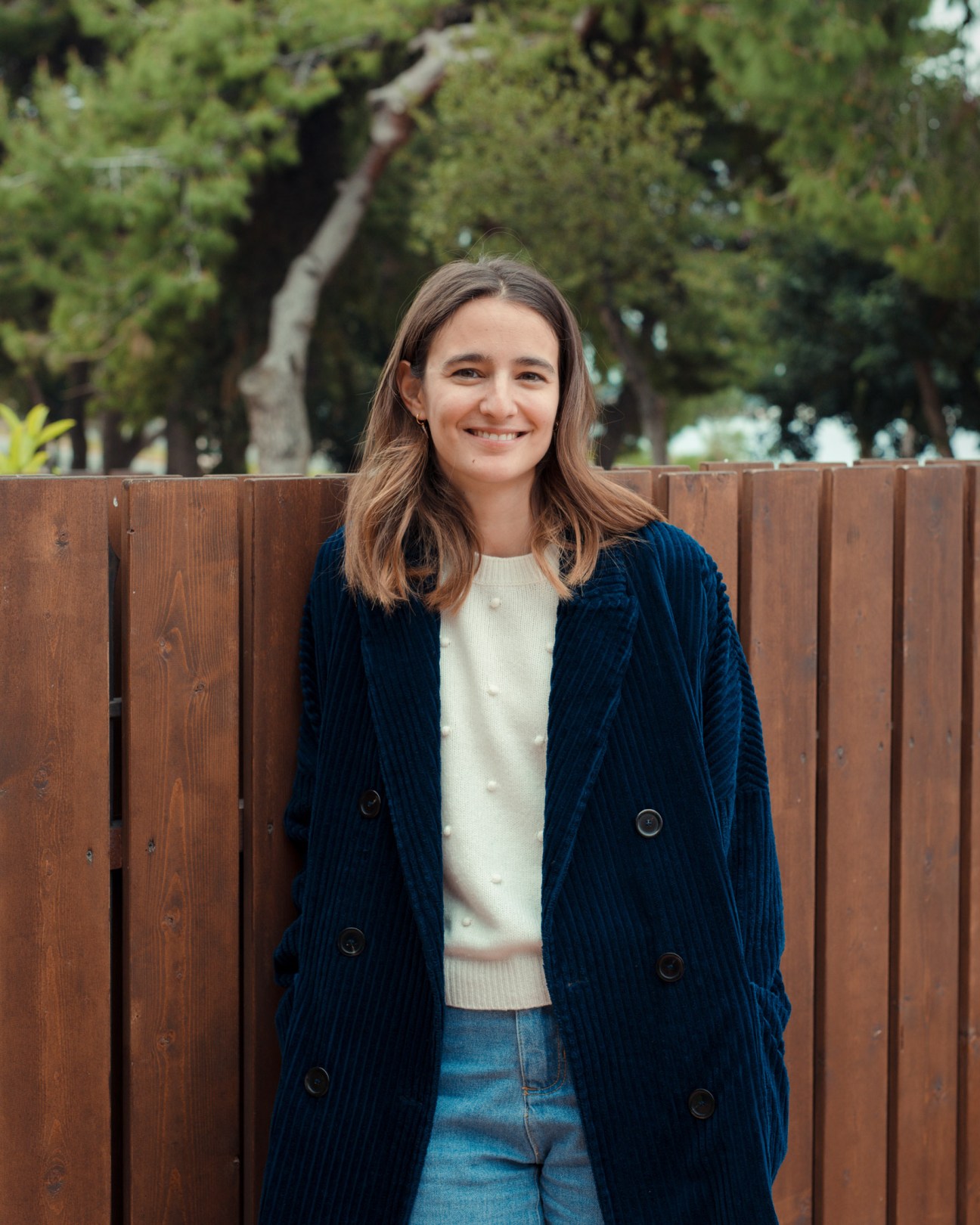 Portrait of Atalanti Martinou, a 32-year-old art curator, smiling against a wooden fence with trees in the background.
