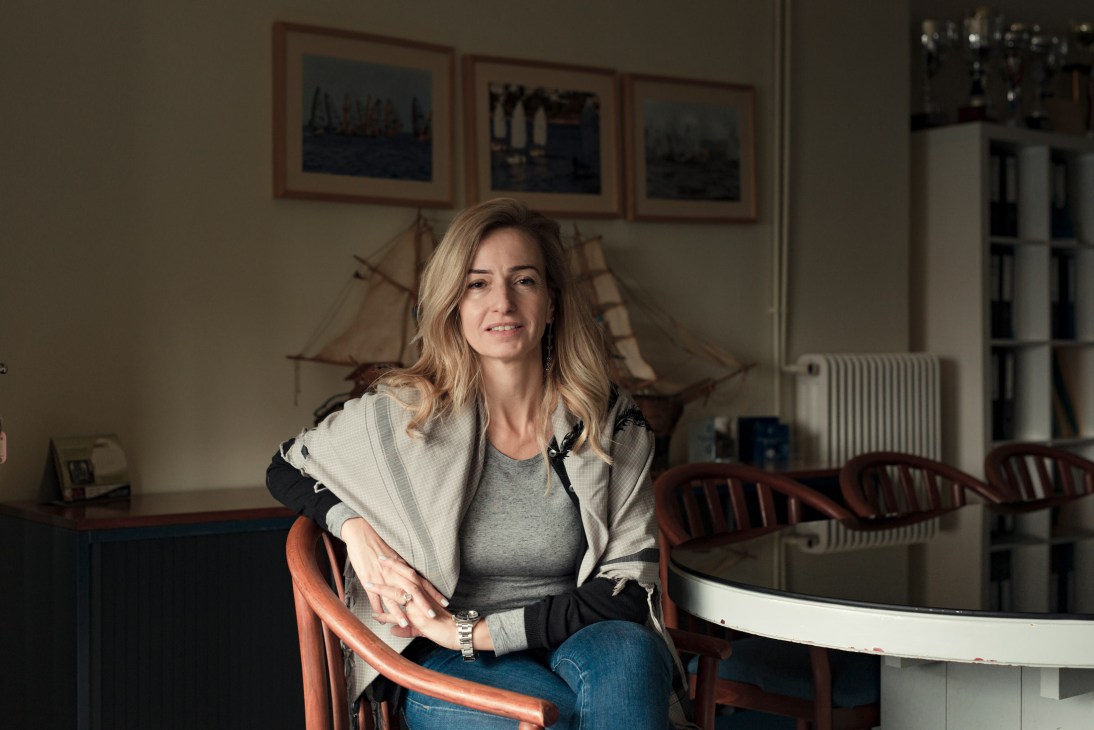 Woman seated indoors with framed maritime artwork and model ship visible on wall behind her.