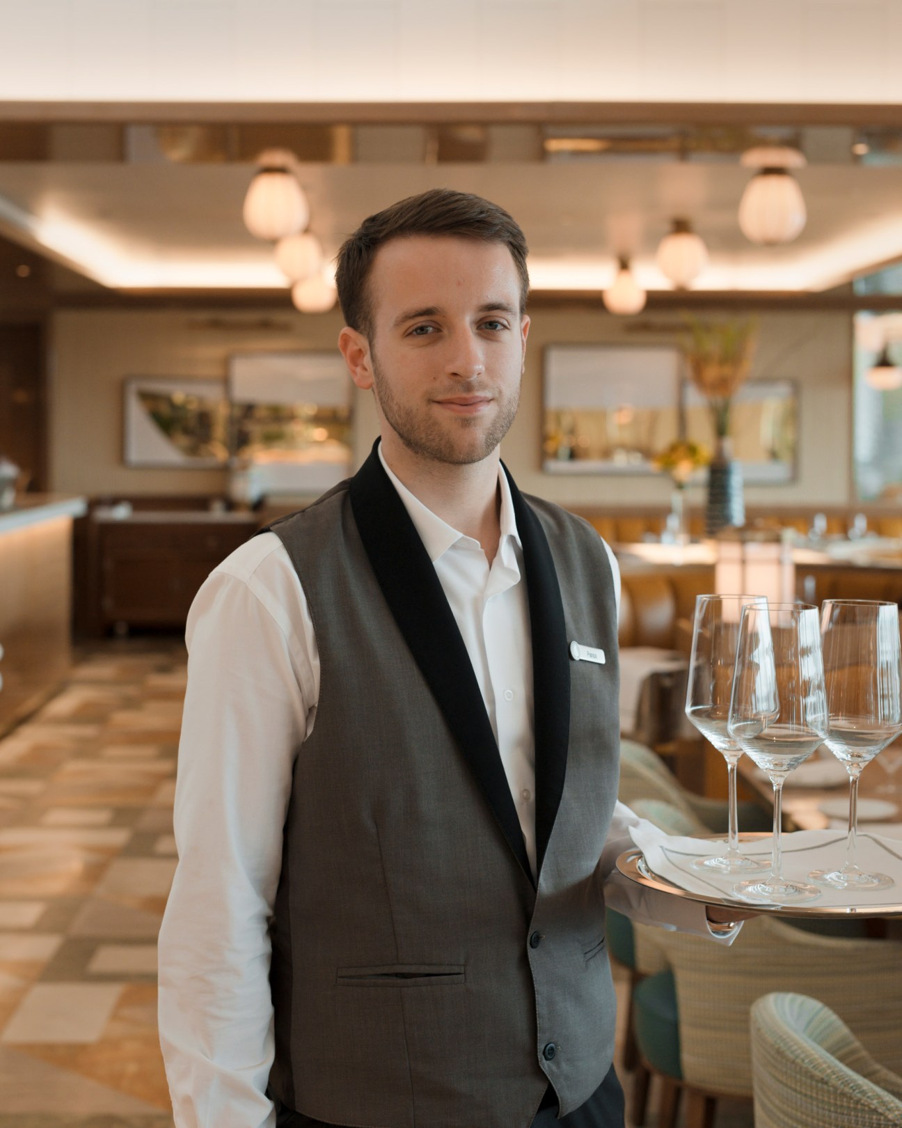Male hotel staff member in formal attire standing in an elegant dining room with place settings.