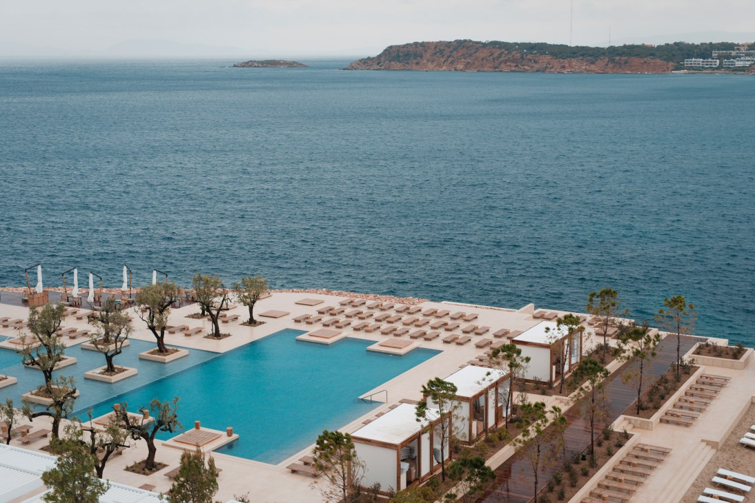 Aerial view of Four Seasons Astir Palace Hotel with turquoise pool, sunbeds, and Aegean Sea coastline with red cliffs beyond.