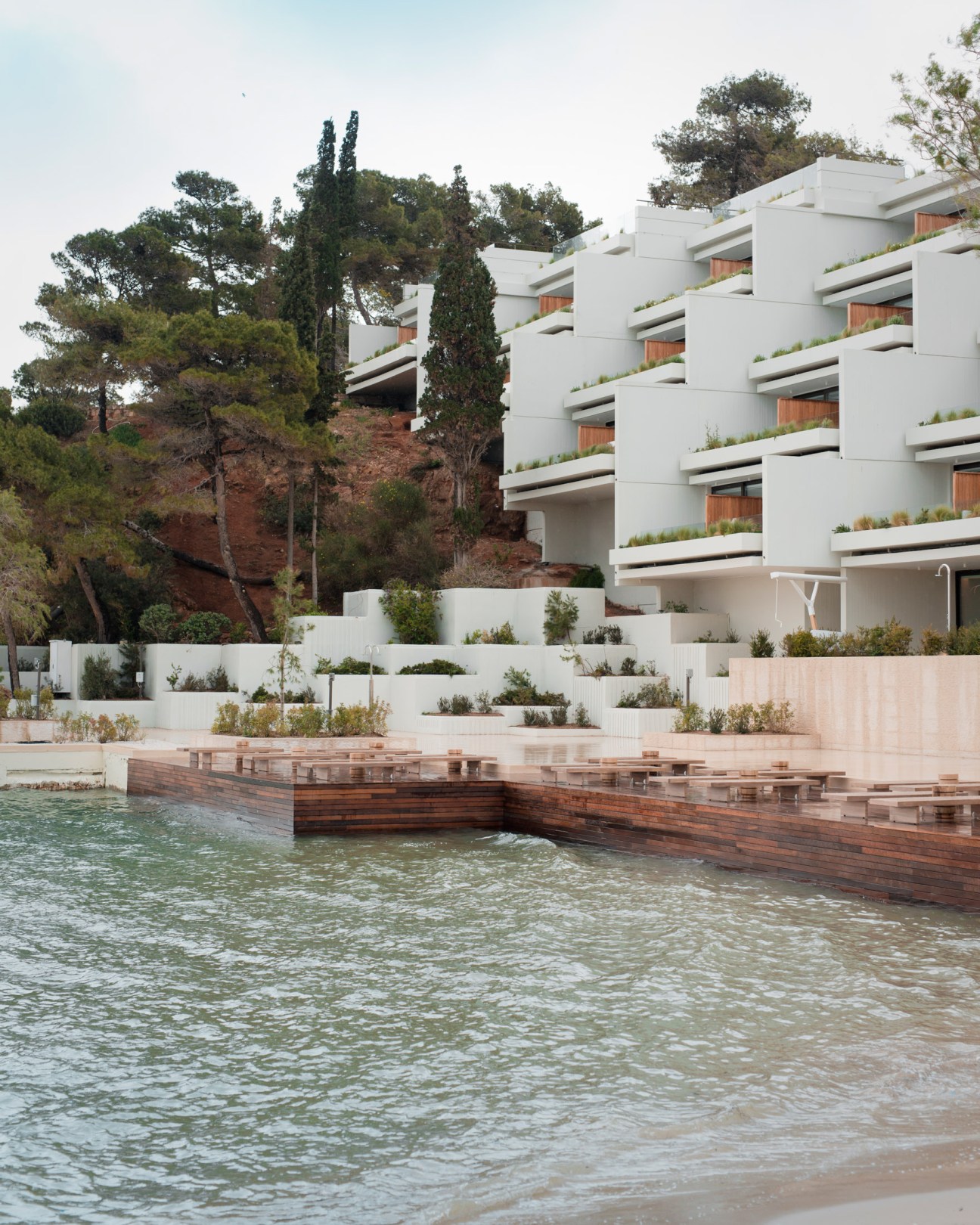 Modernist white apartment building with horizontal balconies overlooking a wooden boardwalk and Aegean Sea.