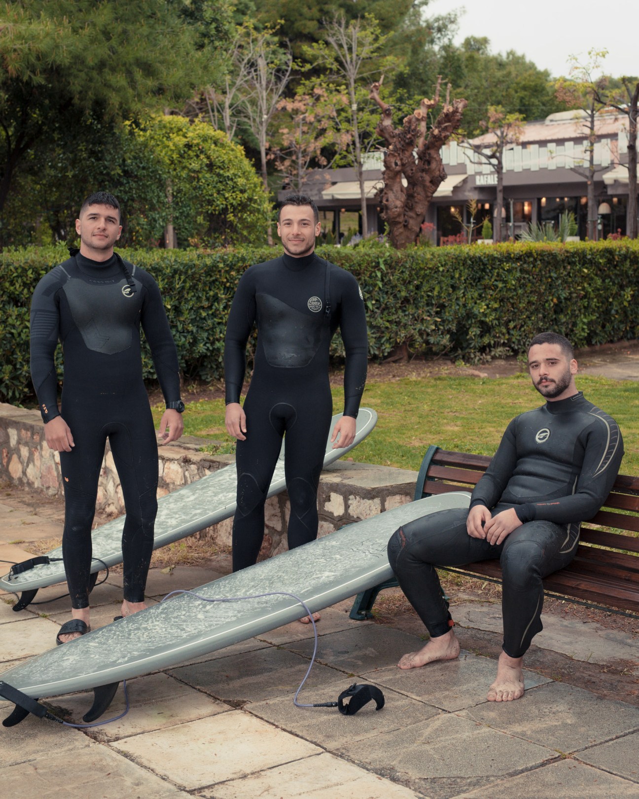 Three surfers in wetsuits rest on a bench with surfboards after watersports at Vouliagmeni.