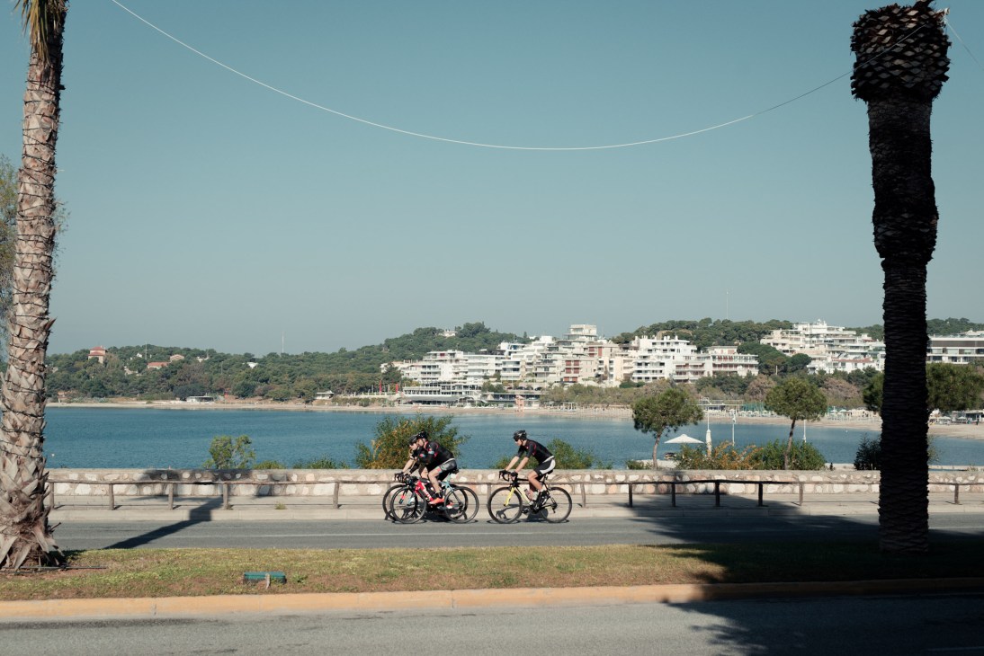Two cyclists ride along a waterfront promenade with palm trees and a coastal view of Vouliagmeni bay and beachfront buildings.