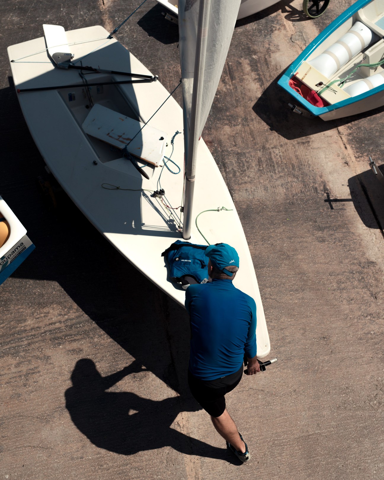 Overhead view of a sailor in blue shirt preparing a small white sailboat at a dock.
