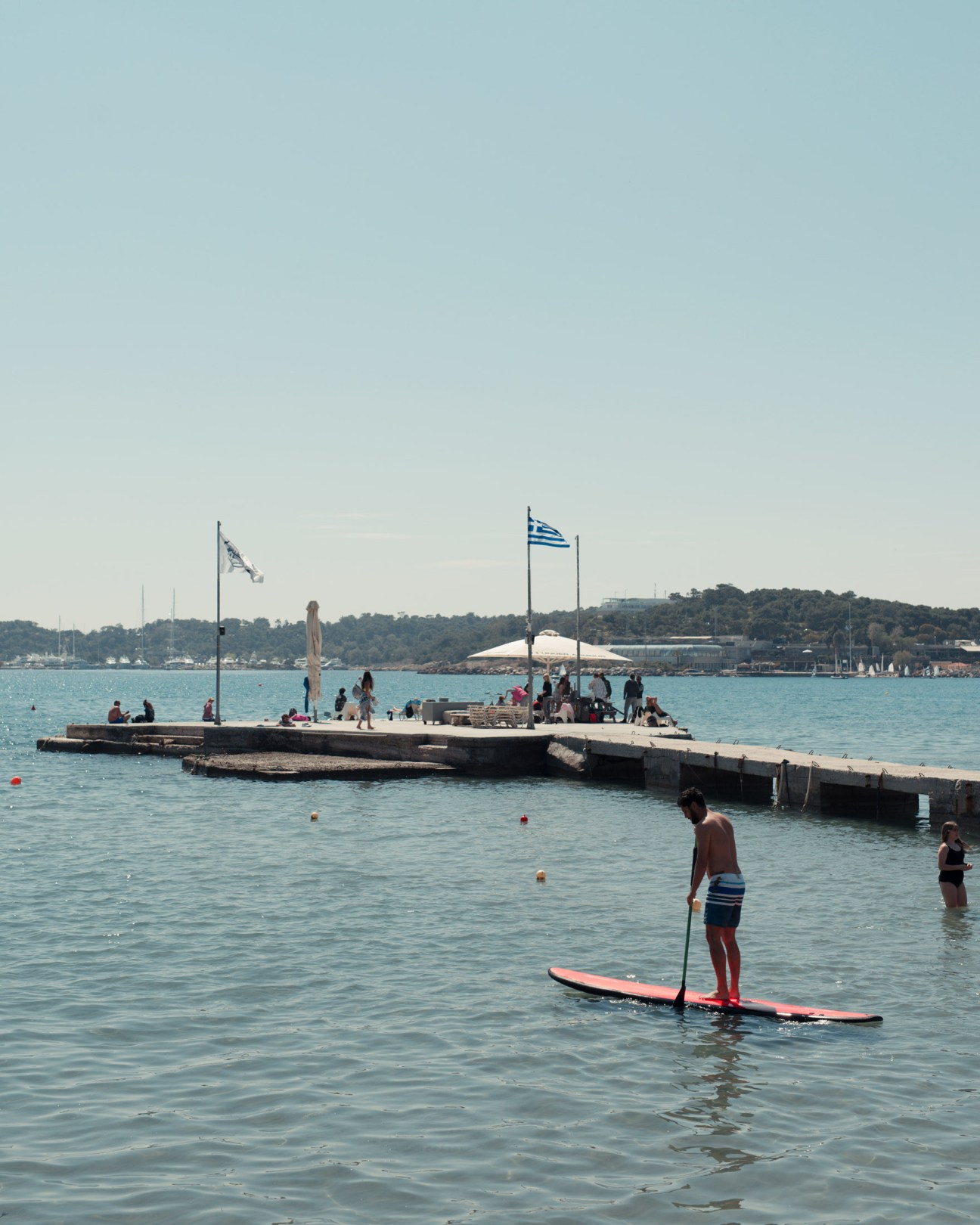 A man paddle boards in turquoise water near a concrete jetty with Greek flags where swimmers and people gather.