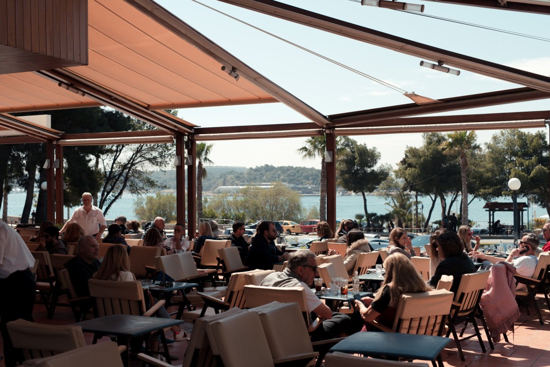 Outdoor café terrace with diners under retractable awnings overlooking the Aegean Sea and forested coastline.