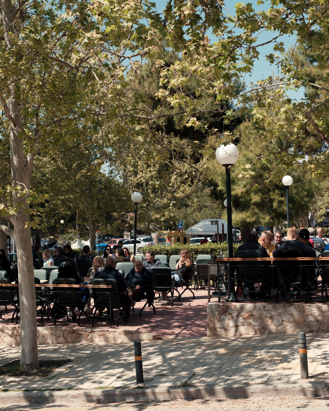 Tree-lined Mediterranean street with outdoor seating and locals gathering in Vouliagmeni