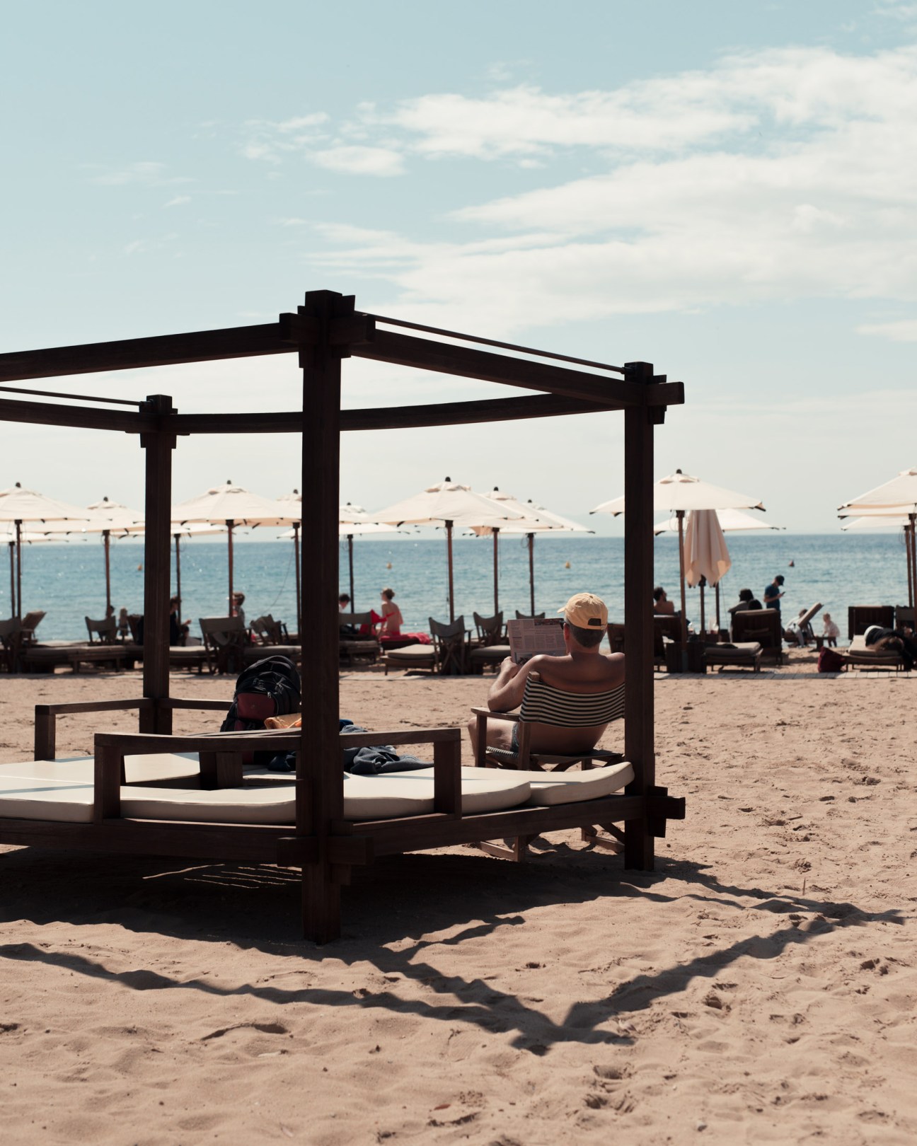 A beachgoer sits beneath a wooden pergola on sand at Astir Beach, with umbrellas and sunbathers visible along the shoreline.