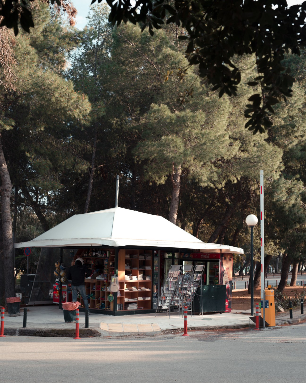 A white-canopied kiosk beneath tall pine trees on a tree-lined Vouliagmeni street.